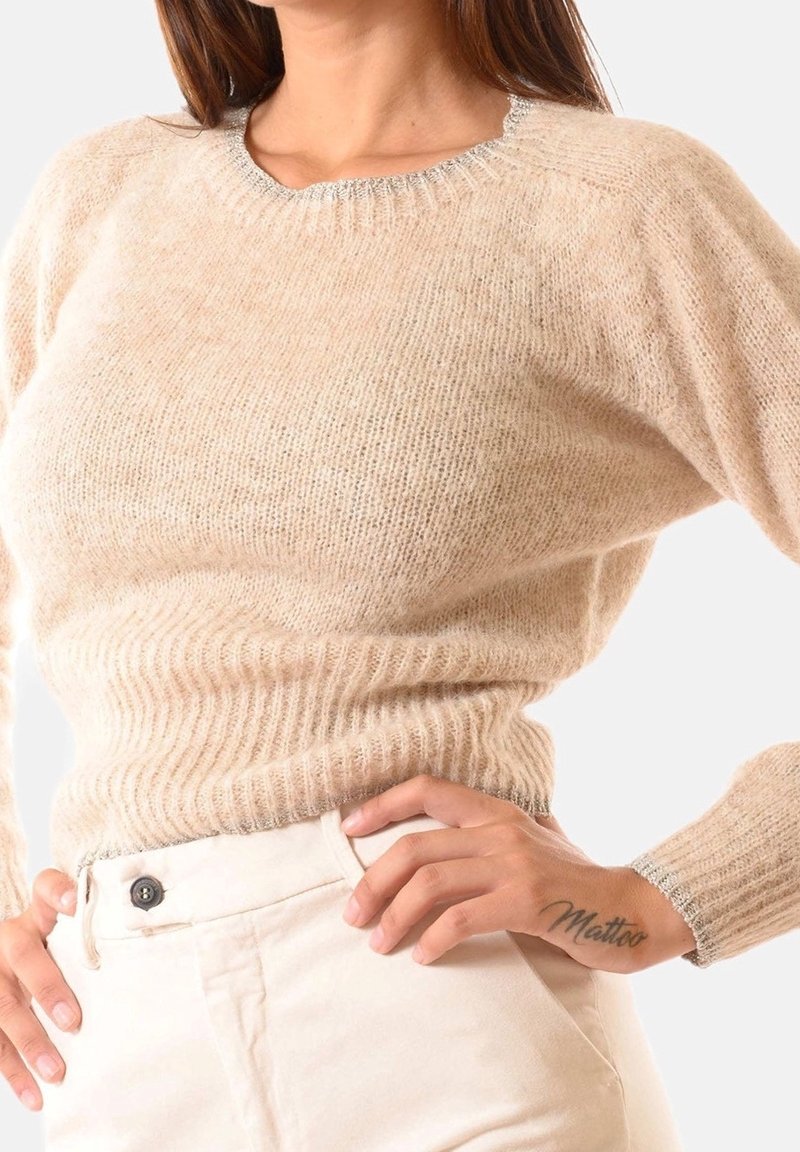 Femme portant un pull en maille beige duveteuse avec un bord argenté, associé à un pantalon crème taille haute, mains sur les hanches dévoilant un tatouage sur la main.