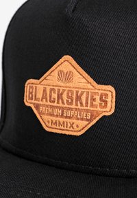 Fekete sapka texturált anyagból, barna bőr folttal. A folton a "BLACKSKIES PREMIUM SUPPLIES MMIX" felirat látható gravírozott betűkkel.
