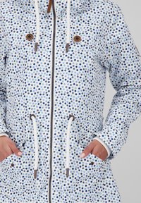 Wodoodporna kurtka o białym tle, zdobiona kropkowanym wzorem w różnych odcieniach niebieskiego. Posiada kaptur, sznurki ściągające oraz duże kieszenie.