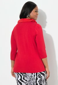 Frau mit langen, dunklen Haaren, die ein rotes Oberteil mit drei Viertel Ärmeln und schwarze-weiße gemusterte Hose trägt, seitlich gegen eine weiße Wand posiert.