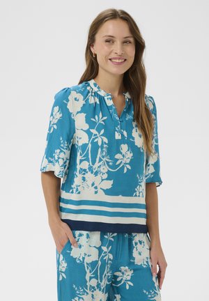 Kobieta uśmiechająca się, ubrana w niebieską kwiecistą bluzkę z rękawami do łokcia i pasujące spodnie, stojąca z rękami w kieszeniach na jednolitym tle.