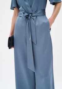 Blaue Textilhaut mit einem Wickeldesign und gebundenem Taillenbereich. Verfügt über weite Ärmel und eine schwarze Clutch, die in einer Hand gehalten wird.