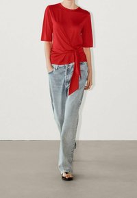 Top rosso a maniche corte con dettaglio annodato in vita, abbinato a jeans a gamba larga azzurri. Tessuto morbido; vestibilità casual e rilassata.