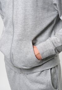 Harmaa vetoketjullinen huppari, valmistettu pehmeästä kankaasta, jossa on sivutaskut ja resorit hihansuuissa. Mallissa on sileä pinta ja rento istuvuus.