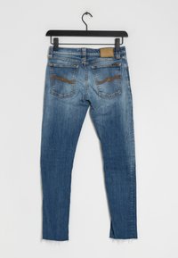 Jeans de mezclilla azul con un acabado deslavado, diseño de cinco bolsillos, acentos de costura marrón y dobladillo sin terminar en los tobillos. Estilo: ajuste ceñido.