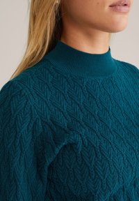 Teal knit sweater with textured herringbone pattern and ribbed collar. Close-up view highlights intricate stitching and soft fabric.