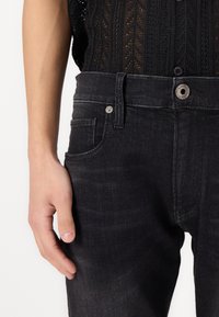 Black denim jeans with a fitted design. Features a button closure, front pockets, and a subtle texture. Seam details along the sides.