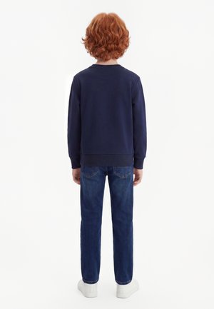 Kind met rood krullend haar, gekleed in een navy trui, blauwe spijkerbroek en witte sneakers, staand met de rug naar voren tegen een effen achtergrond.