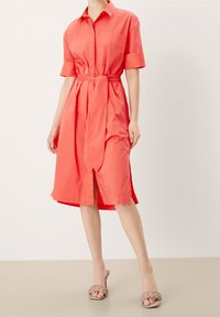 Abito camicia corallo con maniche corte, vita aderente con cintura, gonna a lunghezza ginocchio, chiusura frontale con bottoni e spacchi laterali. Indossato con sandali neutri.