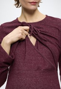 Bordowa bluzka z długim rękawem wykonana z teksturowanego materiału, z dekoltem w kształcie litery V i wiązaniem na środku. Tkanina ma subtelny melanżowy wzór.