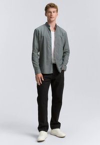 Camicia da uomo grigio chiaro con bottoni, maniche lunghe, taschino sul petto, abbinata a pantaloni neri e sneakers bianche. Tessuto liscio, vestibilità casual.