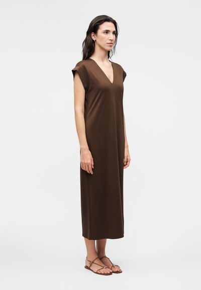 Γυναίκα στέκεται φορώντας ένα καφέ μαν sleeveless φόρεμα μέχρι τους αστραγάλους με V-λαιμόκοψη και ταμπά φλατ σανδάλια μπροστά σε απλό ανοιχτό φόντο.