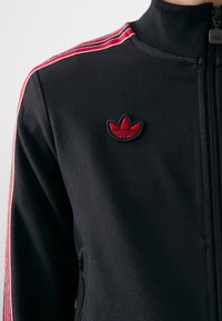 Črna tekaška jakna iz teksturirane tkanine, z rdečimi in rožnatimi črtastimi poudarki, visokim ovratnikom in vezenim rdečim in črnim logotipom.