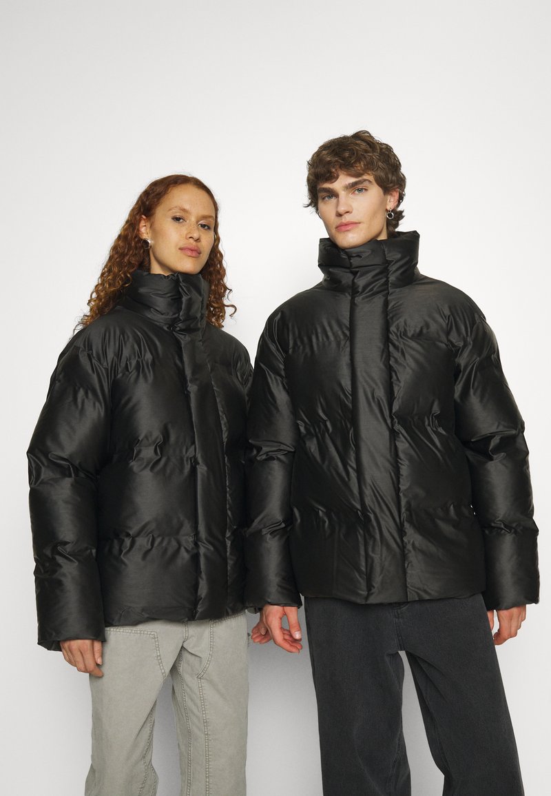 Chaquetas de plumas negras con cuellos altos, que presentan una textura brillante, secciones cosidas para aislamiento, llevadas sobre jeans grises y oscuros.