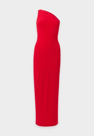 Femme aux cheveux bouclés portant une robe rouge asymétrique avec une fente sur la cuisse et des bottes noires montantes à talons, debout devant un fond uni.