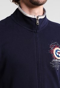 Chaqueta de cremallera azul marino con un alto collar, forro interior gris y un estampado gráfico que presenta acentos rojos y beige en el pecho.