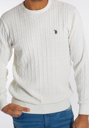 Mann trägt weißen Pullover mit Zopfmuster und Logo auf der Brust sowie blaue Jeans, eine Hand in der Tasche, vor einfarbigem Hintergrund.