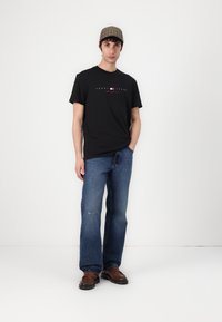 Schwarzes Baumwoll-T-Shirt mit Logo-Print, kombiniert mit blauen Jeans und braunen Slippern. Die Kappe hat ein kariertes Muster.