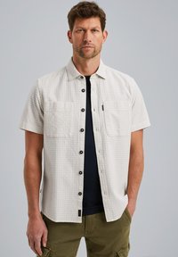 Camisa de manga corta, de color beige claro, con un patrón texturizado, dos bolsillos frontales y botones oscuros. Se lleva sobre una camiseta de color azul marino.