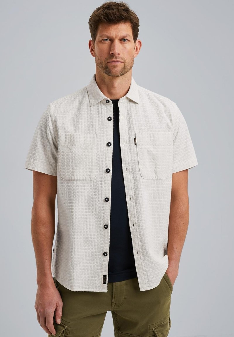 Camisa de manga corta, de color beige claro, con un patrón texturizado, dos bolsillos frontales y botones oscuros. Se lleva sobre una camiseta de color azul marino.