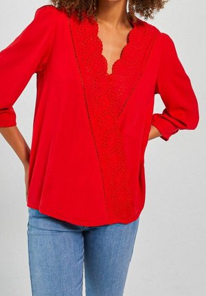 Mujer con blusa roja brillante con diseño de escote en V con bordes festoneados y perforaciones, y mangas tres cuartos, combinada con jeans azul claro.