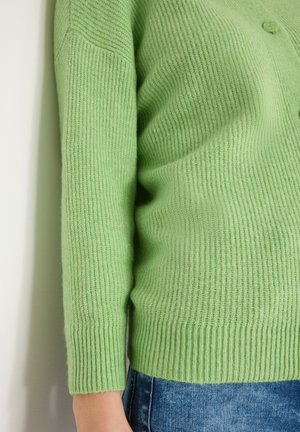 Grön stickad cardigan med ribbad textur, knappstängning och figursydda ärmar, kombinerad med blå jeans, mot en vit bakgrund.