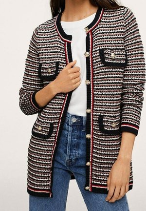 Femme portant un long cardigan texturé à rayures avec des boutons dorés, quatre poches avant, une chemise blanche et un jean bleu, debout.