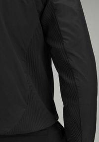 Melna jaka ar gludu audumu un ribotām tekstūras daļām. Strukturēts dizains ar kontrastējošām matētām un spīdīgām virsmām uz piedurknēm.