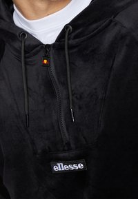 Felpa nera in pile con zip a un quarto, cordini e tasca frontale. Presenta un logo "ellesse" ricamato e una trama sottile.