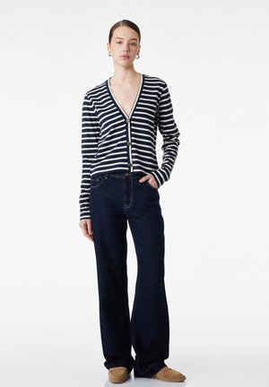 Jonge vrouw draagt een marineblauw-wit gestreepte V-hals cardigan met knopen, donkere wijde jeans en beige suède schoenen, staand met één hand in de zak.