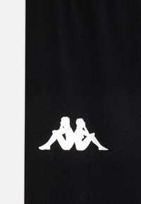 Črno blago krasi velika bela grafika dveh sedečih figur s prekrižanimi nogami, obrobljenih z minimalističnim dizajnom, postavljena na sredino materiala.