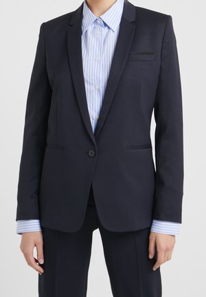 Person iført en mørk marineblazer med en enkelt knap over en lyseblå og hvidstribet skjorte med knapper.