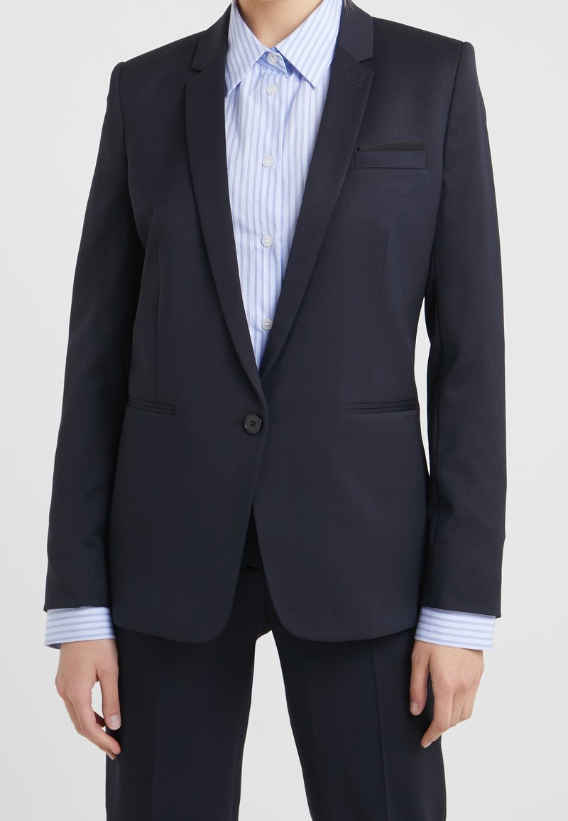 Personne portant un blazer bleu marine foncé à un seul bouton sur une chemise à rayures bleu clair et blanc à boutons.
