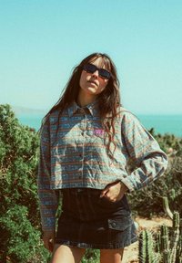 Femme portant des lunettes de soleil, une chemise à manches longues à motifs et une jupe noire se tient à l'extérieur près de cactus avec l'oc éan en arrière-plan.