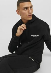 Zwarte hoodie van zachte stof met een voorzak en capuchon met trekkoorden; bevat een witte "Originals"-logo op de borst.