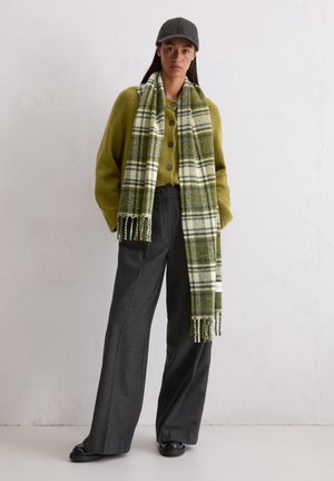 Green plaid scarf with fringed ends draped over an olive green button-up sweater, paired with high-waisted grey trousers and a grey cap.