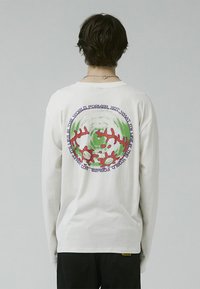 Camiseta de algodón blanca de manga larga con un diseño gráfico colorido en la parte posterior que incluye elementos en verde, rojo y púrpura, además de elementos de texto.
