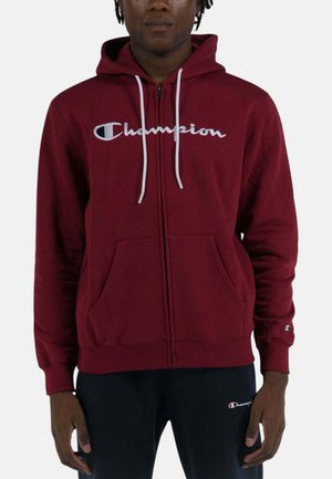 Uomo che indossa una felpa con cappuccio bordeaux Champion con zip e cordini bianchi, e pantaloni neri con il logo Champion.