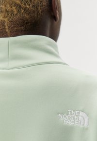 Chaqueta de color verde claro con cuello alto y tejido texturizado. Presenta un logo bordado en blanco en la parte posterior, superficie lisa y un diseño minimalista.