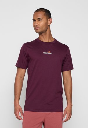 Man wearing a dark purple ellesse t-shirt and muted red shorts, standing against a plain light gray background.