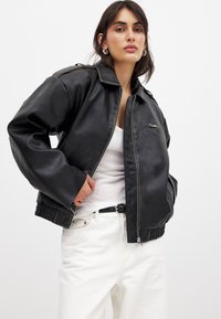 Giacca bomber nera in ecopelle con chiusura a zip, polsini a coste e una finitura testurizzata sottile. Abbinata a una maglia bianca e pantaloni chiari.