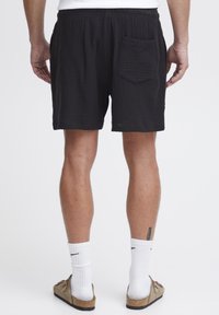 Schwarze Shorts mit strukturierter Oberfläche, weiter Passform und elastischem Bund. Sie haben eine Gesäßtasche und werden mit weißen Socken und Sandalen kombiniert.