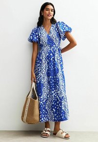 Blaues Blumenkleid mit Puffärmeln, das weiße Blumenmuster und einen Bindegürtel aufweist. Kombiniert mit einer geflochtenen Tasche und flachen Sandalen.