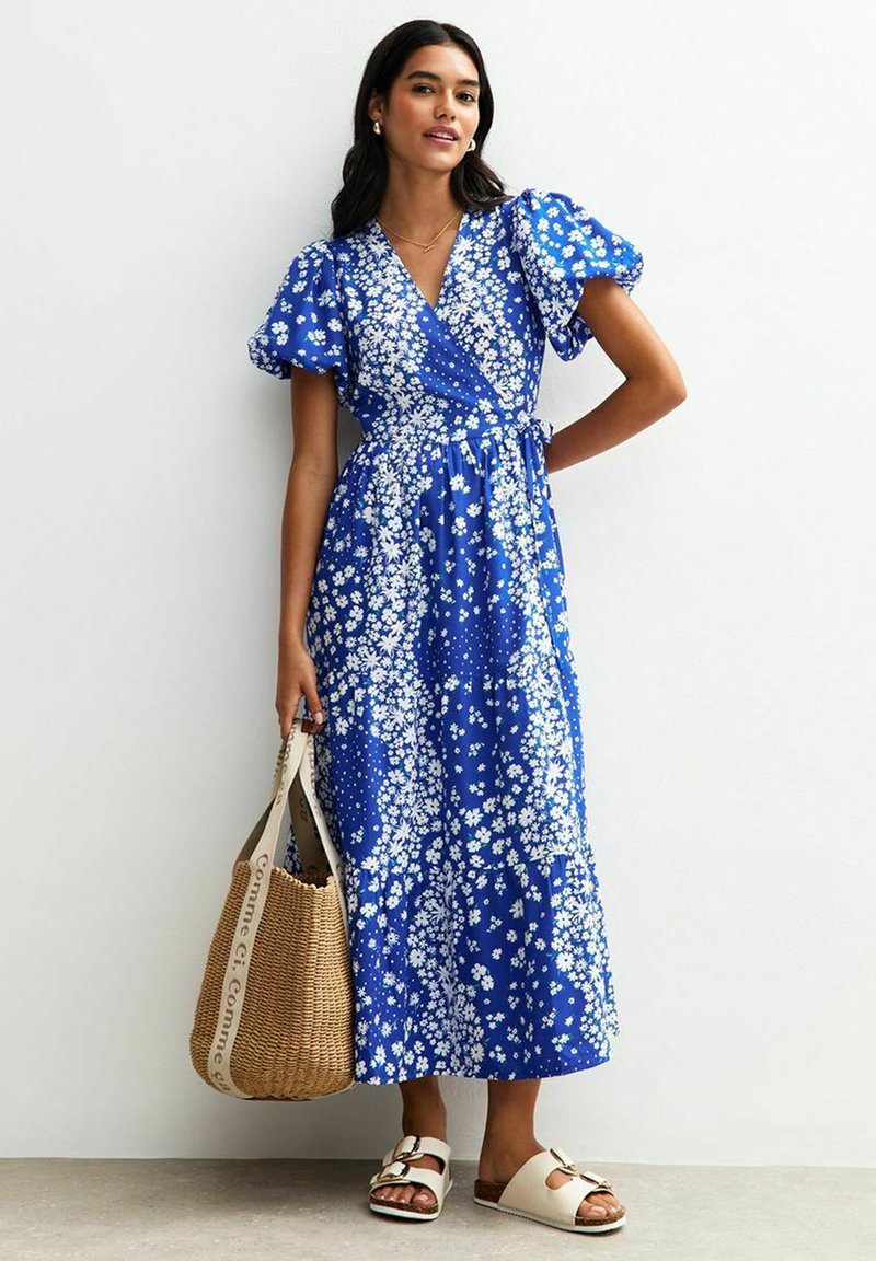 Blaues Blumenkleid mit Puffärmeln, das weiße Blumenmuster und einen Bindegürtel aufweist. Kombiniert mit einer geflochtenen Tasche und flachen Sandalen.