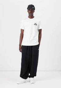 Bílá bavlněná trička s malým modrým logem Nike, zkombinovaná s volnými černými tepláky a bílými teniskami. Černá kšiltovka s bílým logem nahoře.
