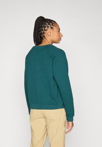 Jūros žalumos spalvos džemperis su apvalia iškirpte ir siaurėjančiomis rankogalių detalėmis, derinamas su kakavos spalvos kelnėmis. Audinys atrodo minkštas ir matinio paviršiaus.