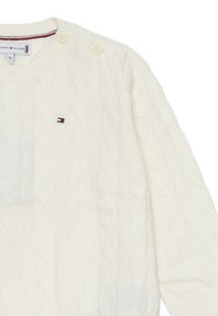 Maglione lavorato a maglia a treccia di colore crema con scollatura rotonda, caratterizzato da due bottoni sulla spalla e un piccolo logo ricamato sul petto.