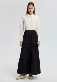Een wit overhemd met een zwarte gelaagde maxi-rok met ruches, gemaakt van een zachte stof, gedragen met lichte schoenen.