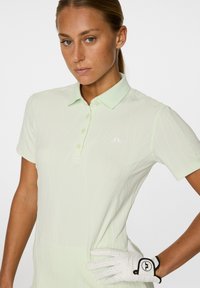 Hellgrünes Golf-Polo mit kurzen Ärmeln, ausgestattet mit einem strukturierten Design, subtiler Glanz und einem Logo auf der Brust. Weißer Golf-Handschuh an der linken Hand getragen.