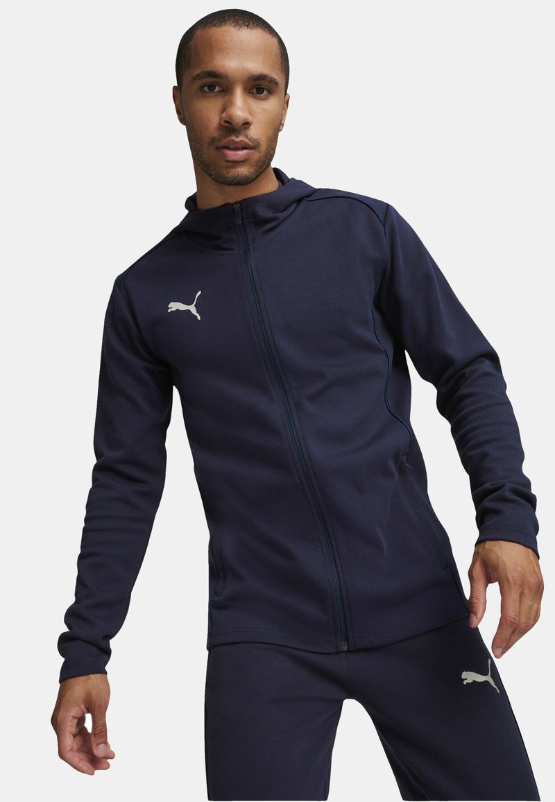 Marineblå zip-up træningsjakke med en tætsiddende pasform. Har en høj krave, lange ærmer og et lyst gråt Puma-logo på venstre bryst.